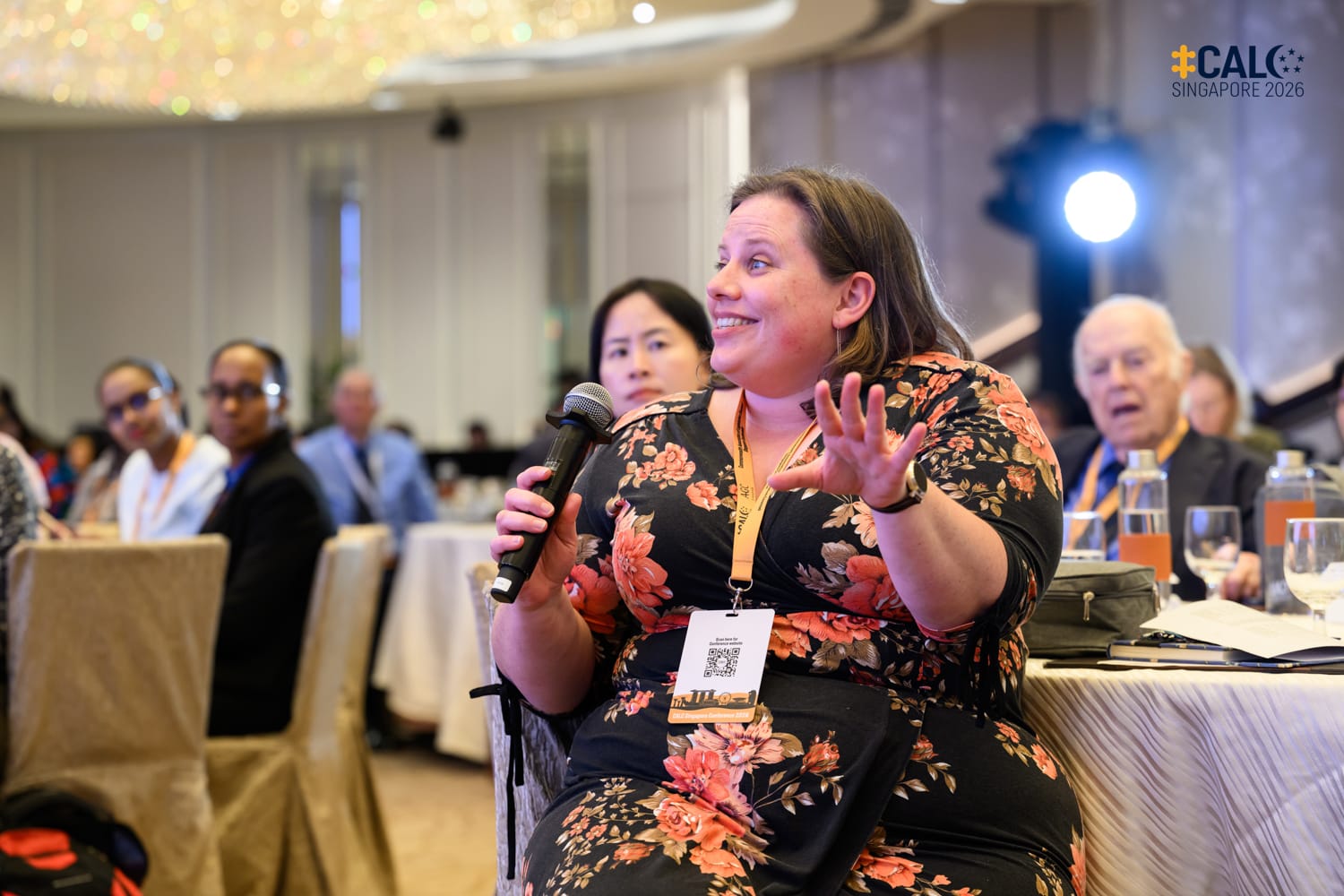 A woman in a floral dress holds a microphone and gestures while speaking at the CALC Singapore 2026 conference, with other attendees seated at tables in the background.